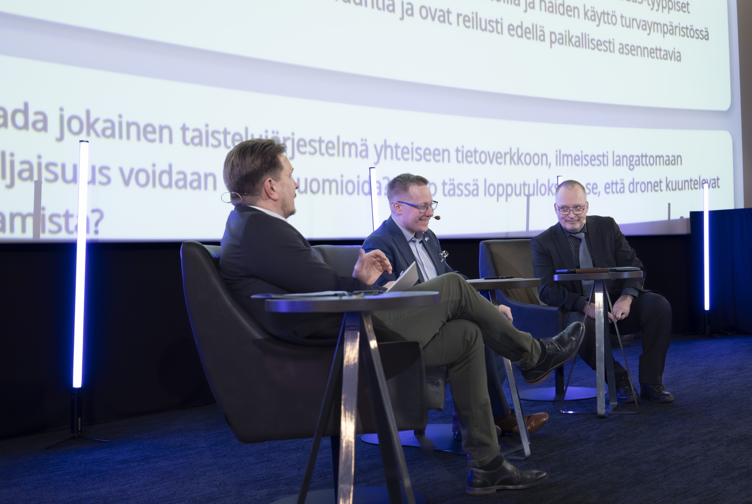 Kolme paneelikeskustelijaa istumassa seminaarissa, jonka aiheena oli puolustusteollisuus. Taustalla yleisön lähettämiä kysymyksiä.