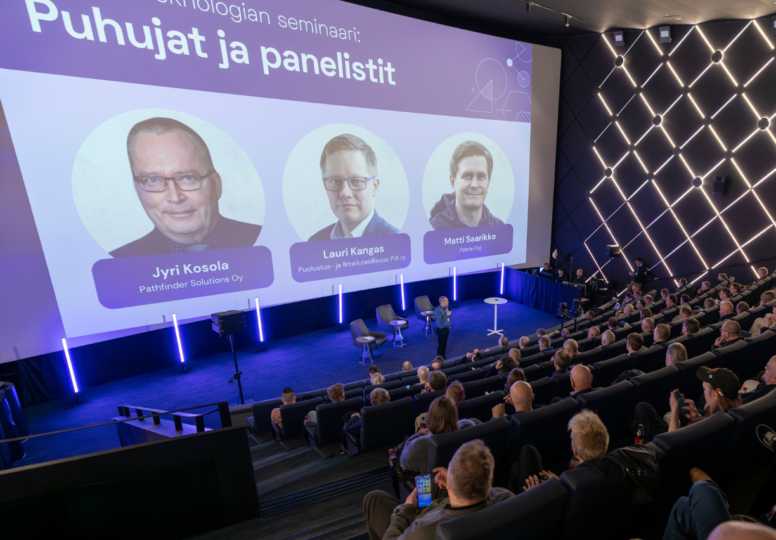 Ihmisiä elokuvateatterissa odottamassa seminaarin alkua, valkokankaalla puhujien kuvat ja nimet.