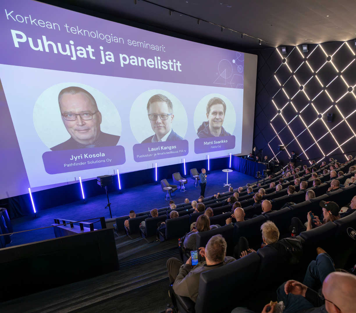 Ihmisiä elokuvateatterissa odottamassa seminaarin alkua, valkokankaalla puhujien kuvat ja nimet.