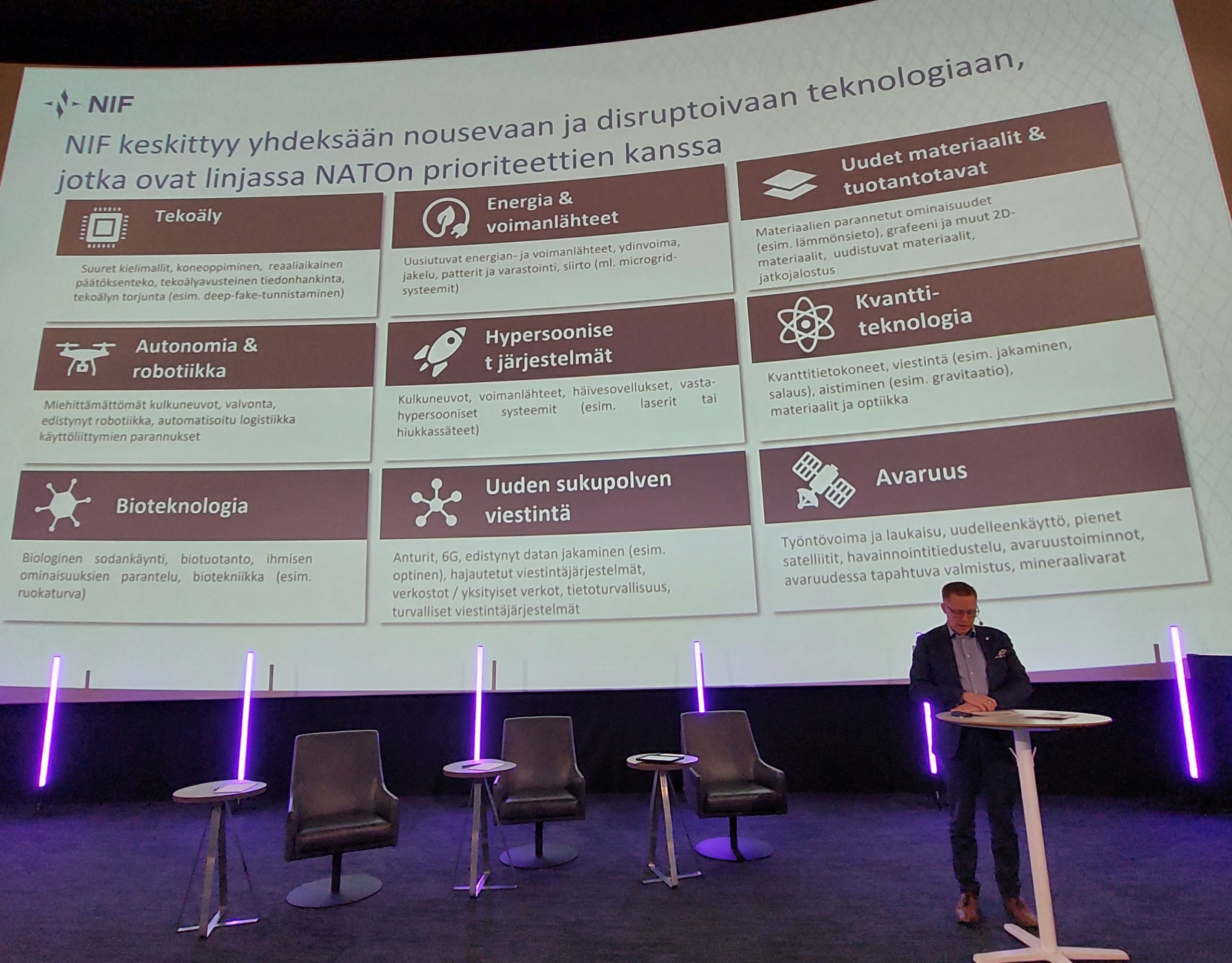 Seminaarin puhuja taustallaan esitysmateriaalia. Aiheena puolustusteollisuus ja sen käyttämät teknologiat.
